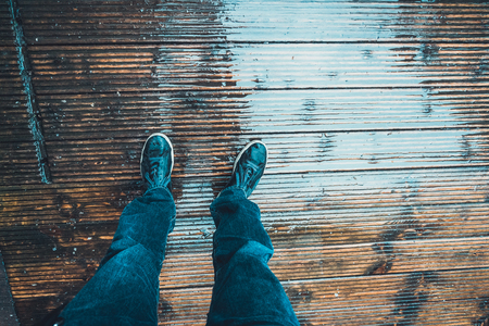 Man standing on a wet wooden deck with a first person perspective looking down at his legs and shoesの写真素材