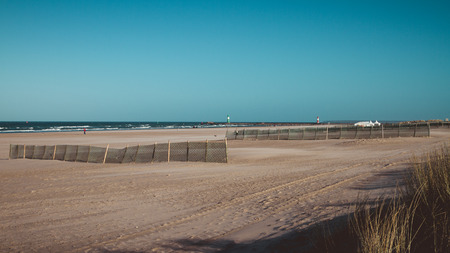 Wide open beach with fences and shadows from grass along ocean shoreline in background under blue skyの写真素材