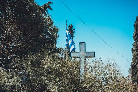 Close Up of Religious Cross and Blue and White Striped Flag of Greece Surrounded by Lush Green Tree Branches on Sunny Day with Clear Blue Sky in Greeceの写真素材