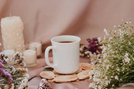 Cup of coffee on a stand surrounded by flowers and candles of different sizes and shapesの写真素材