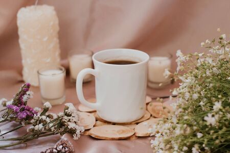 Cup of coffee on a stand surrounded by flowers and candles of different sizes and shapesの写真素材