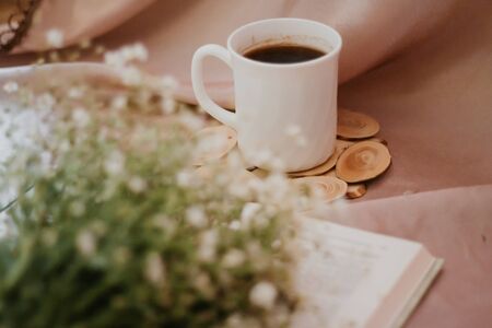 Cup of coffee on a stand surrounded by flowers and bookの写真素材