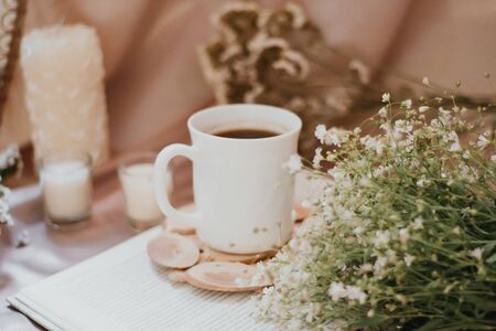 Cup of coffee on a stand surrounded by flowers and candles of different sizes and shapesの写真素材