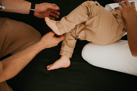 Parents help their toddler adjust pants while seated on a couch, showcasing a cozy home environment with soft textures and neutral colorsの写真素材