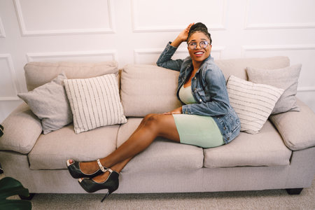 Beautiful young African American woman sitting on sofa and smiling in living roomの写真素材
