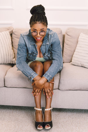 Beautiful young African American woman sitting on sofa and smiling in living roomの写真素材