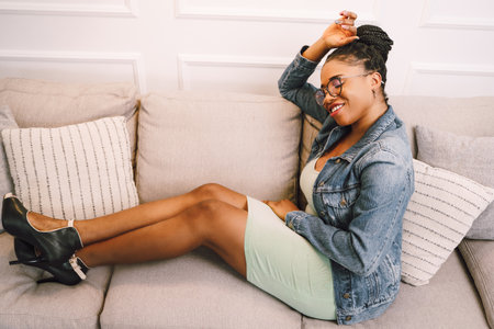 Beautiful young African American woman sitting on sofa and smiling in living roomの写真素材
