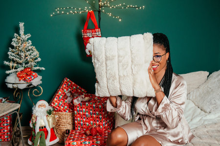 Young happy African American woman in a dress smiling near Christmas tree and playing with pillow on the bedの写真素材