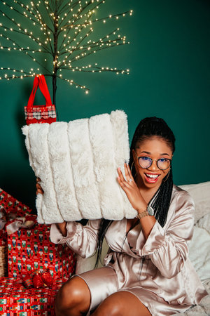 Young happy African American woman in a dress smiling near Christmas tree and playing with pillow on the bedの写真素材
