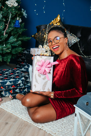 Young beautiful African American woman in red dress sitting and smiling on Christmas decorated backgroundの写真素材