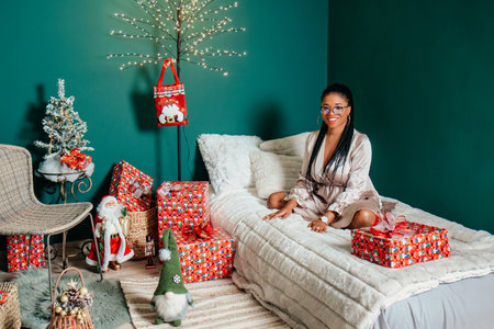 Young happy African American woman in a dress smiling on bed and playing with Christmas decorationの写真素材