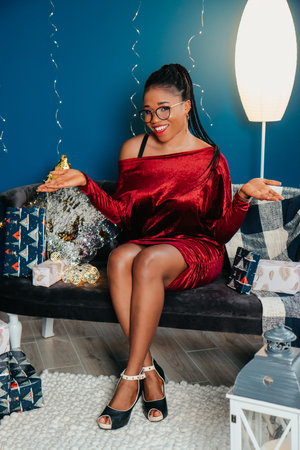 Young beautiful African American woman in red dress sitting and smiling on Christmas decorated backgroundの写真素材
