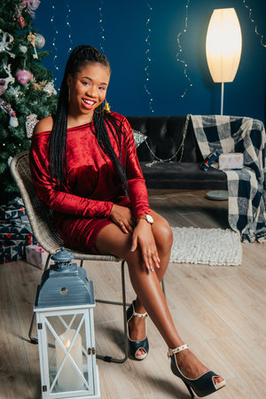 Young beautiful African American woman in red dress sitting and smiling on Christmas decorated backgroundの写真素材