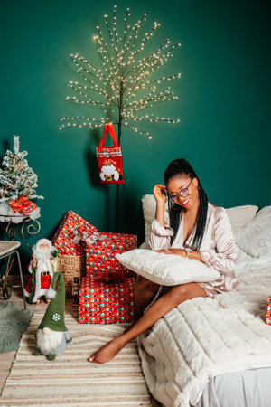 Young happy African American woman in a dress smiling on bed and playing with Christmas decorationの写真素材