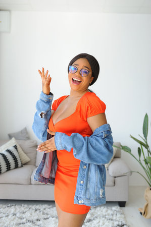 Cheerful young African American woman in orange dress with jacket smiling and dancingの写真素材