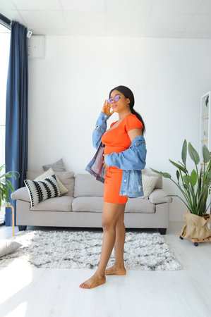 Cheerful young African American woman in orange dress with jacket smiling and dancingの写真素材