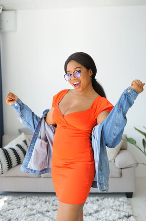 Cheerful young African American woman in orange dress with jacket smiling and dancingの写真素材