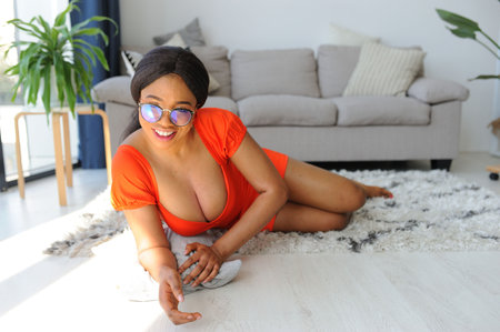 Young beautiful African American woman in an orange dress with jacket smiling and laying down near sofaの写真素材