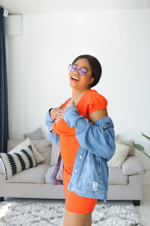 Young beautiful African American woman in an orange dress with jacket smiling and standing near sofaの写真素材