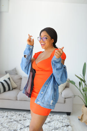 Young beautiful African American woman in an orange dress with jacket smiling and standing near sofaの写真素材