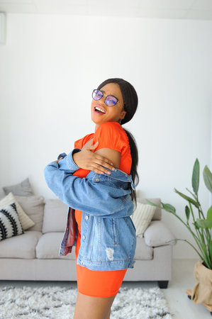 Young beautiful African American woman in an orange dress with jacket smiling and standing near sofaの写真素材