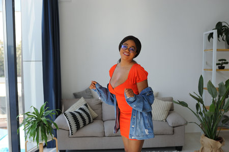 Young beautiful African American woman in an orange dress with jacket smiling and standing near sofaの写真素材
