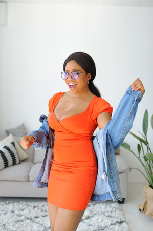 Cheerful young African American woman in orange dress with jacket smiling and dancingの写真素材