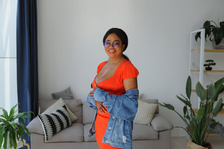 Young beautiful African American woman in an orange dress with jacket smiling and standing near sofaの写真素材