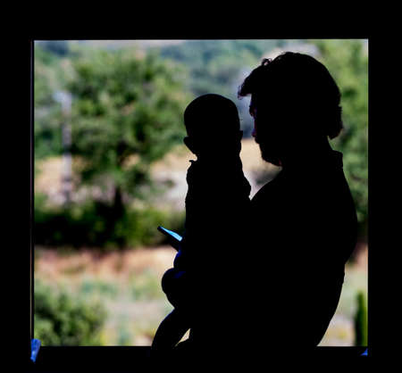 Father looking at smartphone while holding his little child in his arms at the window in silhouette