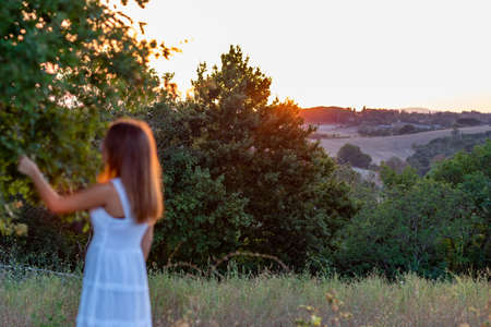 Blurred sunset profile of a young girl with long blonde hair dressed in white as she touches the leaves of the magic treeの写真素材