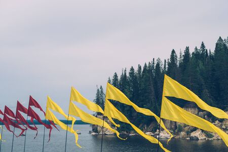 Red and yellow flying flags at rivers bank against rocks, dark forest and grey sky