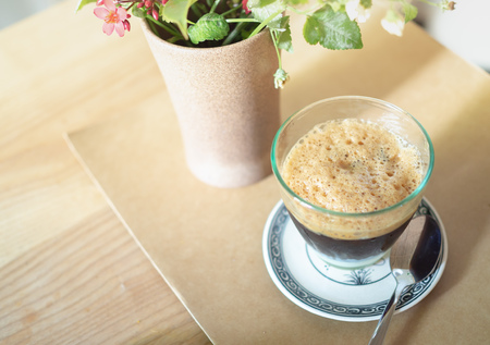 Foam on Top of Coffee with Milk in The Glass with Spoon and Flower in The Vase on Wooden Table for Breakfast in The Morningの写真素材