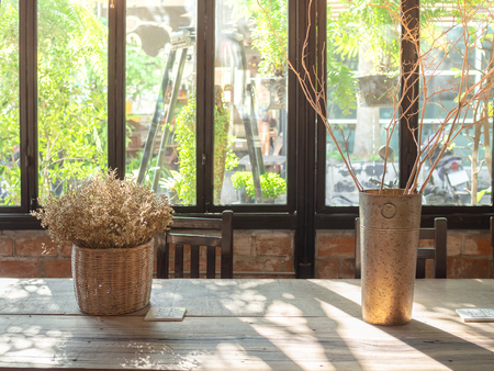 Loft Interior Cafe with Flowers in Vintage Tall Vase, Basket and Plug on Wooden Table with Wooden Chais in front of The Glass Window, Brick Wall and Garden Outside on Sunshine Dayの写真素材