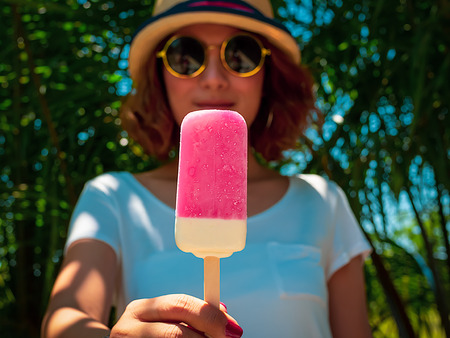 Beautiful Woman Short Hair Wearing White Shirt, Beach Hat and Round Sunglasses with Happy Smiling Holding White and Pink Frozen Ice Pop on Summer Time with Sunshine on Tree and Blue Sky Backgroundの写真素材