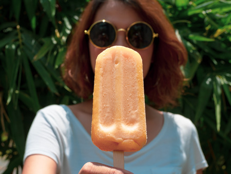 Beautiful Woman Short Hair Wearing White Shirt and Round Sunglasses with Happy Smiling Holding Yellow Frozen  Ice Pop on Summer Time with Sunshine on Tree Backgroundの写真素材