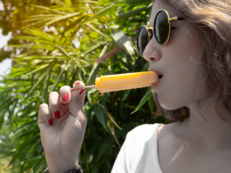 Beautiful Woman Short Hair Wearing White Shirt and Round Sunglasses Eating Frozen Yellow  Ice Pop on Summer Time with Sunshine on Tree Backgroundの写真素材