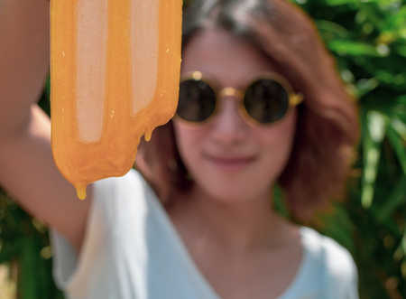 Beautiful Woman Short Hair Wearing White Shirt and Round Sunglasses with Happy Smiling Holding Yellow Bited Ice Pop Upside Down and Melting on Summer Time with Sunshine on Tree Backgroundの写真素材