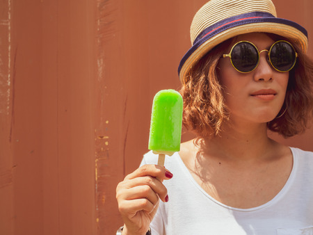 Beautiful Woman Short Hair Wearing White Shirt, Hat and Round Sunglasses with Happy Smiling Holding Green Frozen Ice Pop on Summer Time with Red Container Backgroundの写真素材