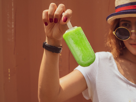 Beautiful Woman Short Hair Wearing White Shirt, Hat and Round Sunglasses with Happy Smiling Holding Green Frozen Ice Pop on Summer Time with Red Container Backgroundの写真素材