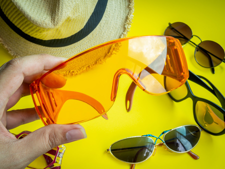 Flat Lay Traveler Accessories. Hand Picking The Orange Summer Plastic Glasses and Many Kind Sunglasses and Beach Hat on Lemon Yellow Background. Top View Vacation Summer Concept.の写真素材
