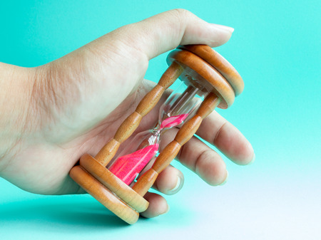 Hand Holding The Wooden Vintage Hourglass with Pink Sand on Blue Background, Sand Running through the Bulbs for Measuring The Passing Time in A Countdown to Deadline.の写真素材
