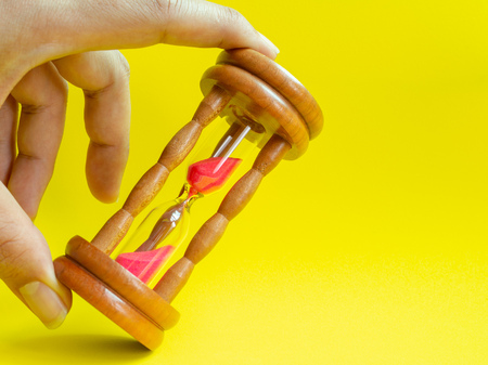 Hands Holding The Wooden Vintage Hourglass with Pink Sand on Yellow Background, Sand Running through the Bulbs for Measuring The Passing Time in A Countdown to Deadline.の写真素材