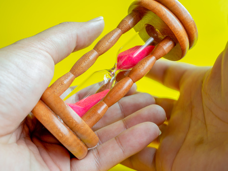 Hands Holding The Wooden Vintage Hourglass with Pink Sand on Yellow Background, Sand Running through the Bulbs for Measuring The Passing Time in A Countdown to Deadline.の写真素材