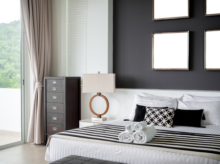 Modern Bedroom with Four Square Blank Stainless Steel Frames on The Gray Wall Interior, Pillows and Towels on The Bed with Side Lamp and Wooden Vintage Cabinet Near The Glass Sliding Door.の写真素材