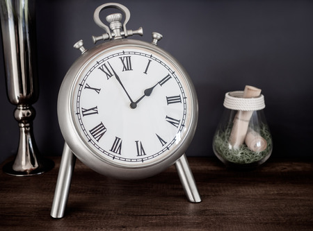Close-up Stainless Classic Vintage Roman Numerals Clock, Glass and Vase. Still Life on Wooden Cabinet on Dark Background.の写真素材
