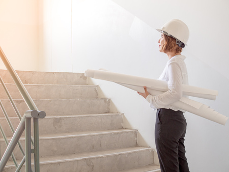 Beautiful asian woman engineer curly short hair wearing white safety helmet and white shirt holding blueprint on Stair on wall construction building background. Working woman concept.の写真素材