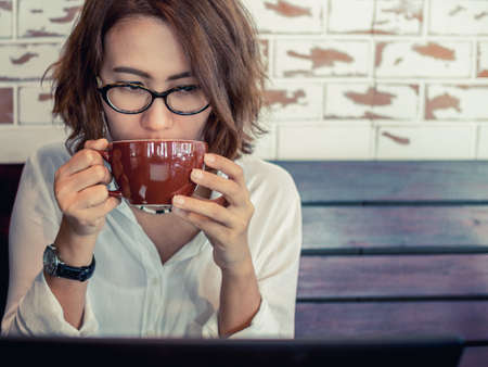 Beautiful Asian Woman wearing glasses and white shirt drinking coffee and watching to her black laptop screen on white brick wall and wooden background in cafe.の写真素材