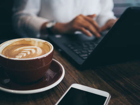 Hand typing on Black laptop computer with computer mouse, concentrated at working with coffee and mobile phone on wooden table in cafe. Working woman concept.の写真素材