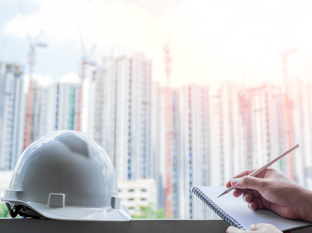 White hard safety helmet for civil engineer, protect and safety hat for engineering worker and hand writing notebook by pencil on construction building site background.の写真素材