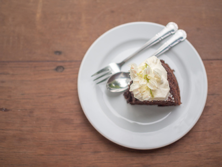 A piece of wild organic chocolate honey cake with beautiful vintage flowers topping and whipping cream on top in white ceramic plate with sillver spoon and fork on wooden table background top view with copy space.の写真素材